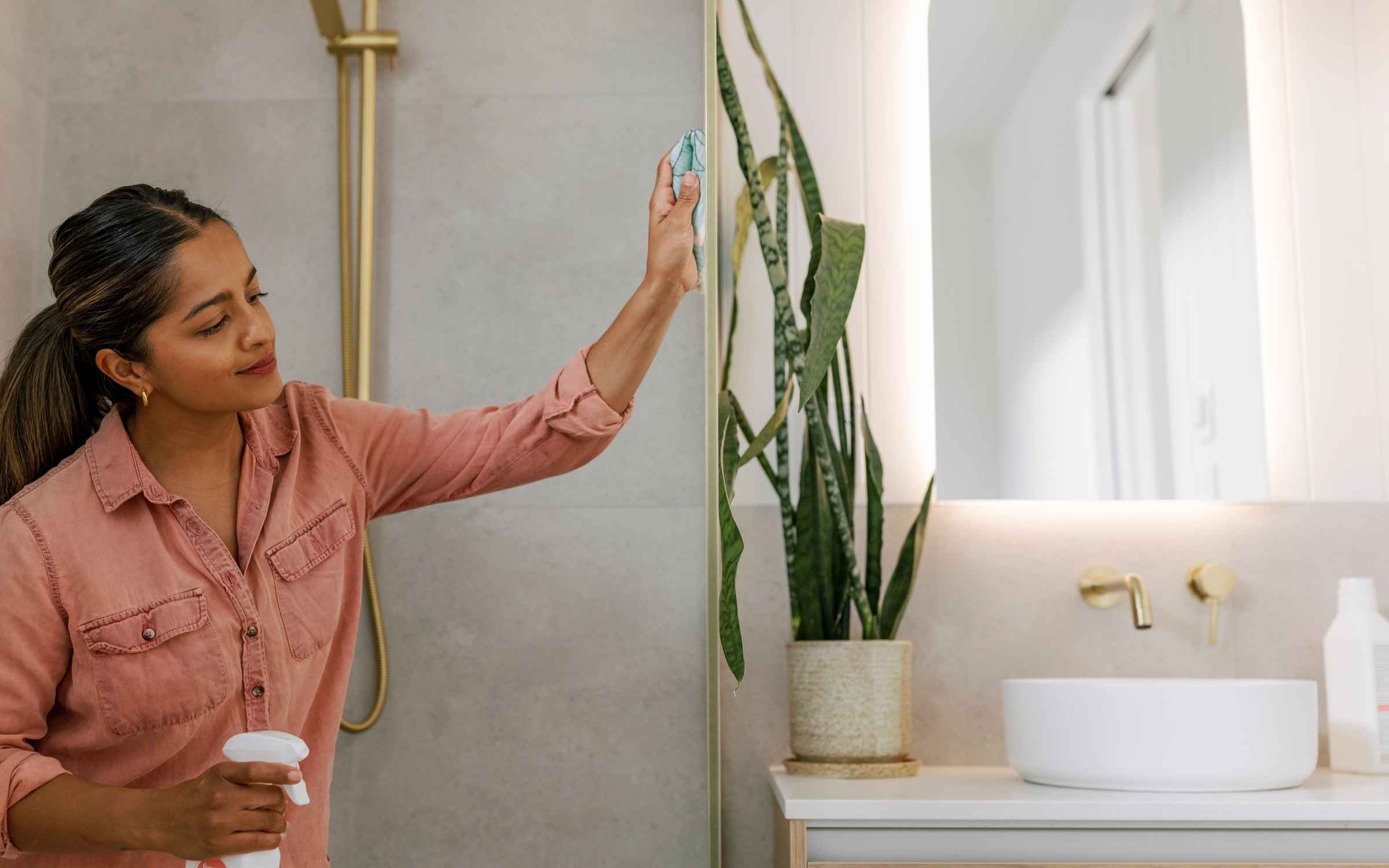 Woman wiping down a shower glass panel with a Forsana spray bottle in hand, in a bright modern bathroom